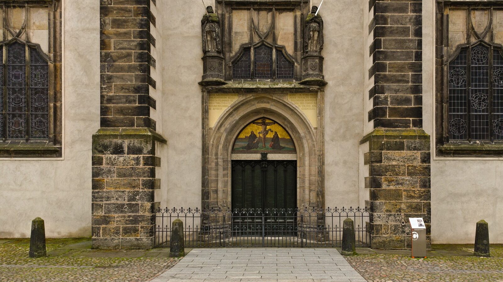 Fachada da Igreja do Castelo de Wittenberg, na Alemanha, com a Porta das Teses de bronze sob um arco gótico e um mosaico da crucificação no alto, entre dois contrafortes de pedra.